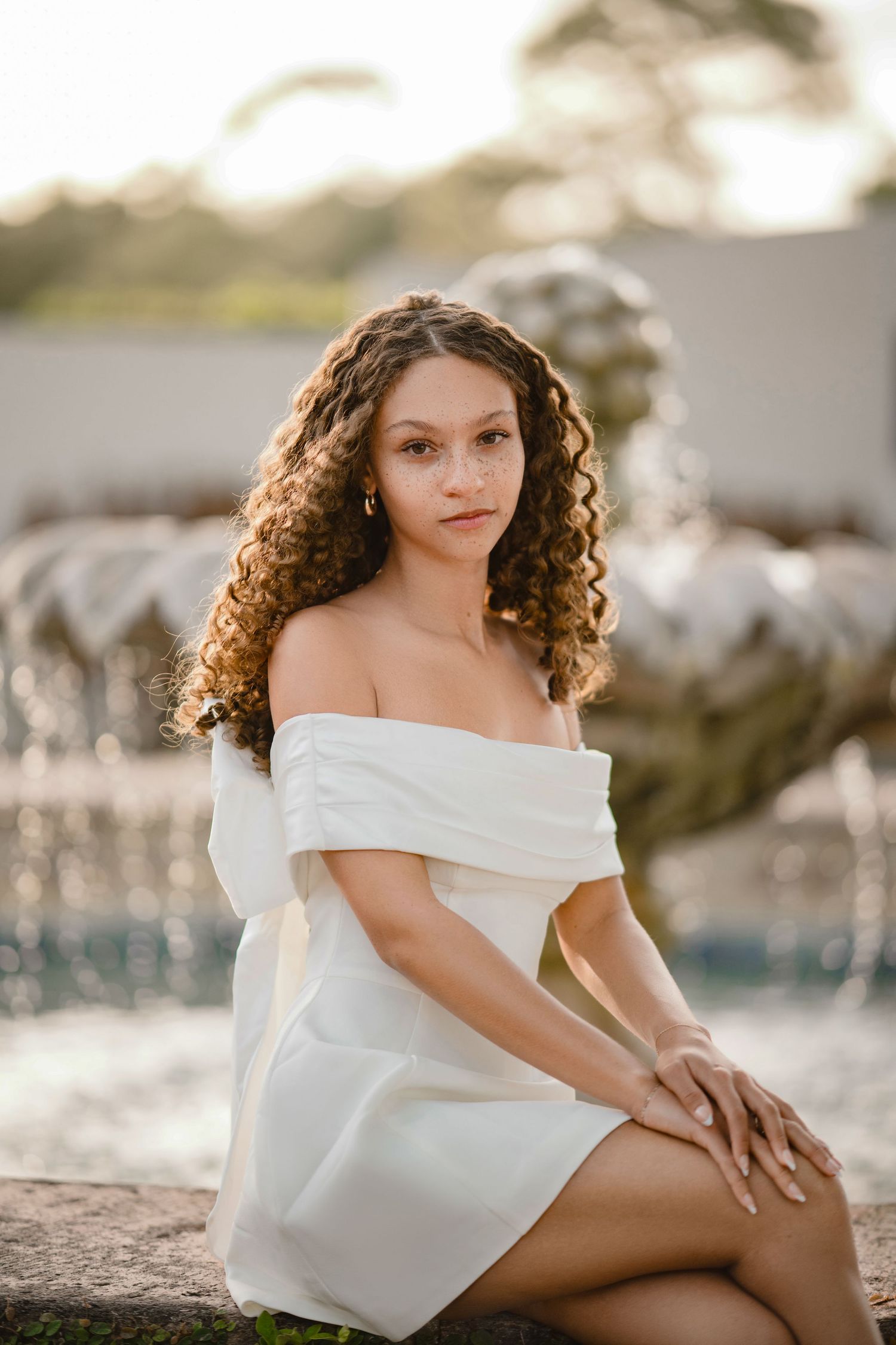 Young woman with long curly hair sits on a stone ledge in a white off-the-shoulder dress in front of a softly blurred fountain at sunset.