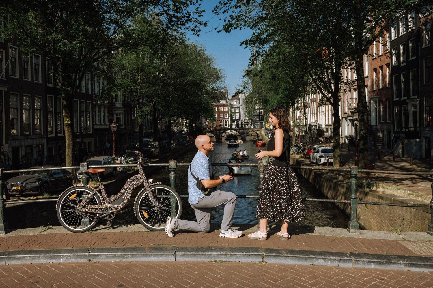 Man kneeling on one knee proposing to a woman in a dress beside a tree-lined canal in Amsterdam, with a bicycle parked nearby.