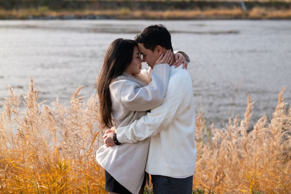A romantic couple in white tops embraces tenderly among golden tall grass beside a calm lake at sunset.