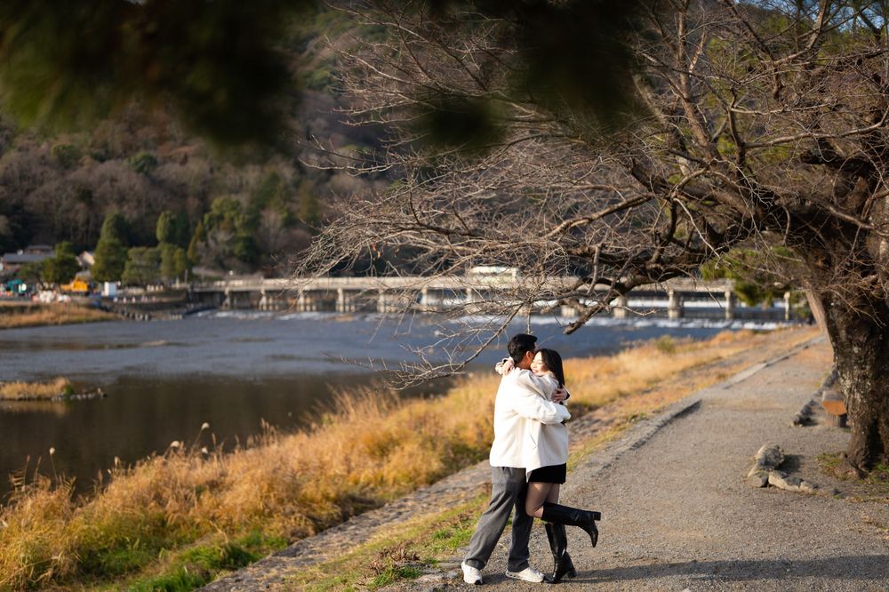 Couple embracing and kissing on a scenic riverside path with golden grass, bare trees, and a bridge in the background.