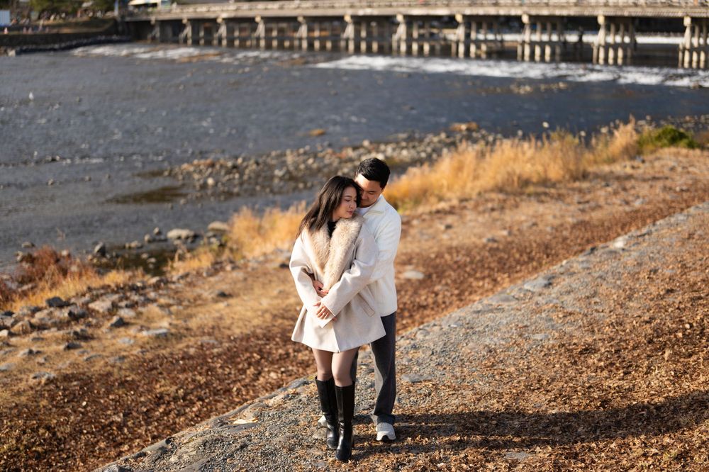 A couple in white coats embraces on a sunlit, leaf-covered riverbank with a long bridge in the background.