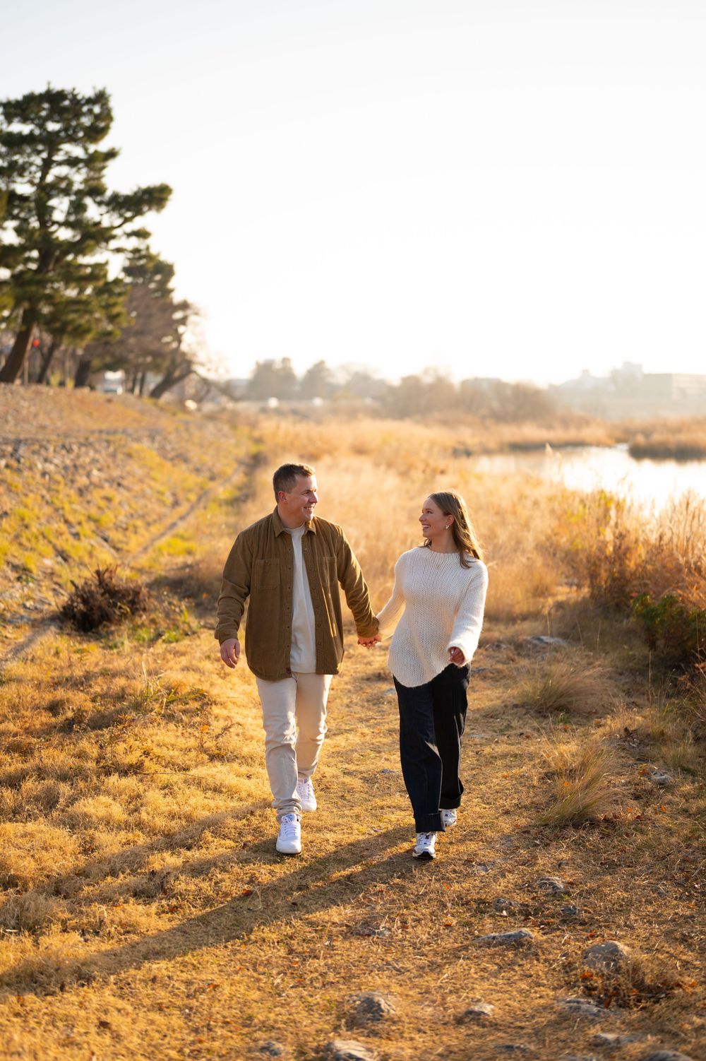 Couple holds hands while walking along a dirt path through golden grass beside a calm lake at sunset.