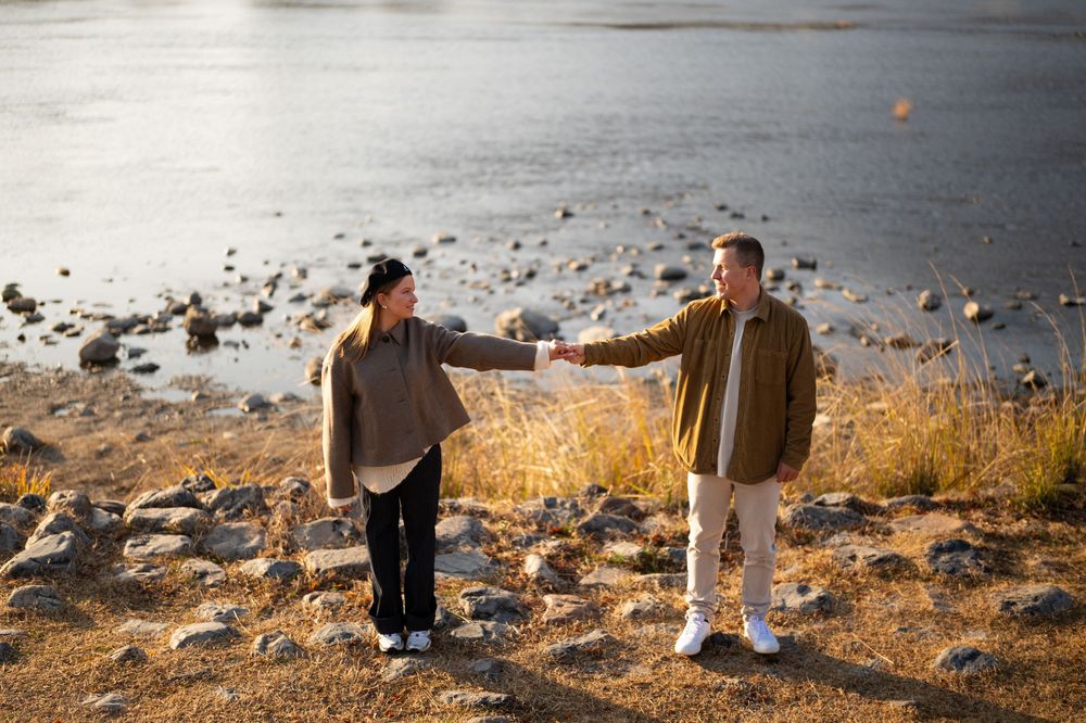 Two people in casual jackets hold hands while walking across a rocky lakeshore at golden hour, with calm water and dry grass behind them.