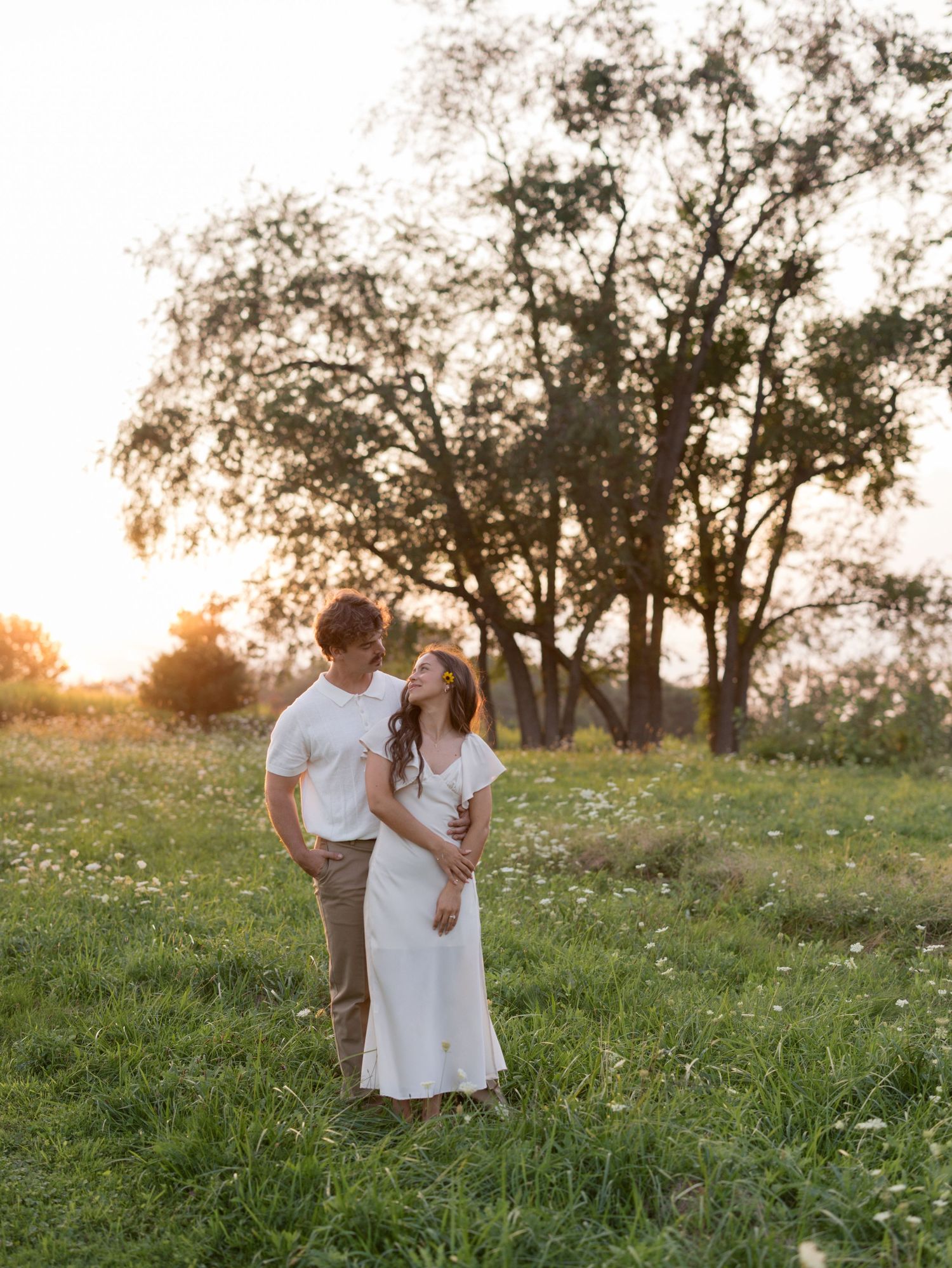Couple embracing in a green meadow with wildflowers at golden hour sunset, framed by tall trees in a romantic outdoor engagement photo.