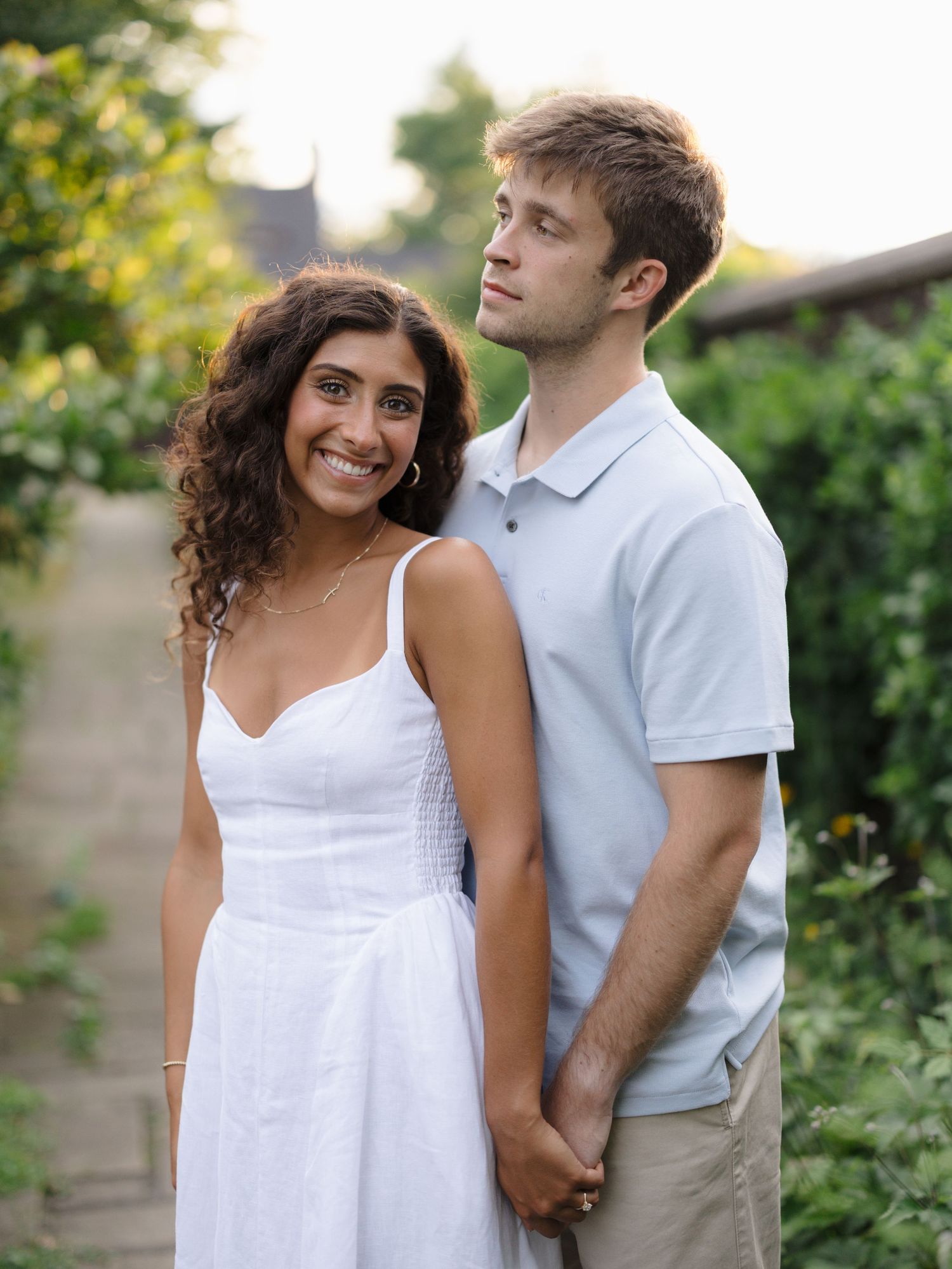 Smiling woman in a white sundress holds hands with a man in a light blue polo as they stand together in a sunlit garden path.
