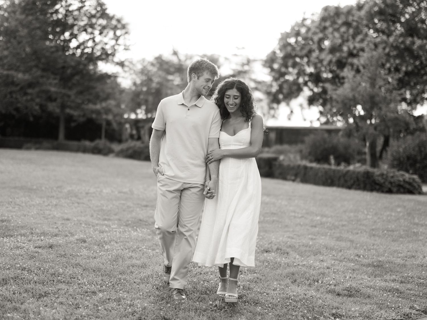 Black and white photo of a smiling couple holding hands and walking across a grassy lawn, with trees in the background.