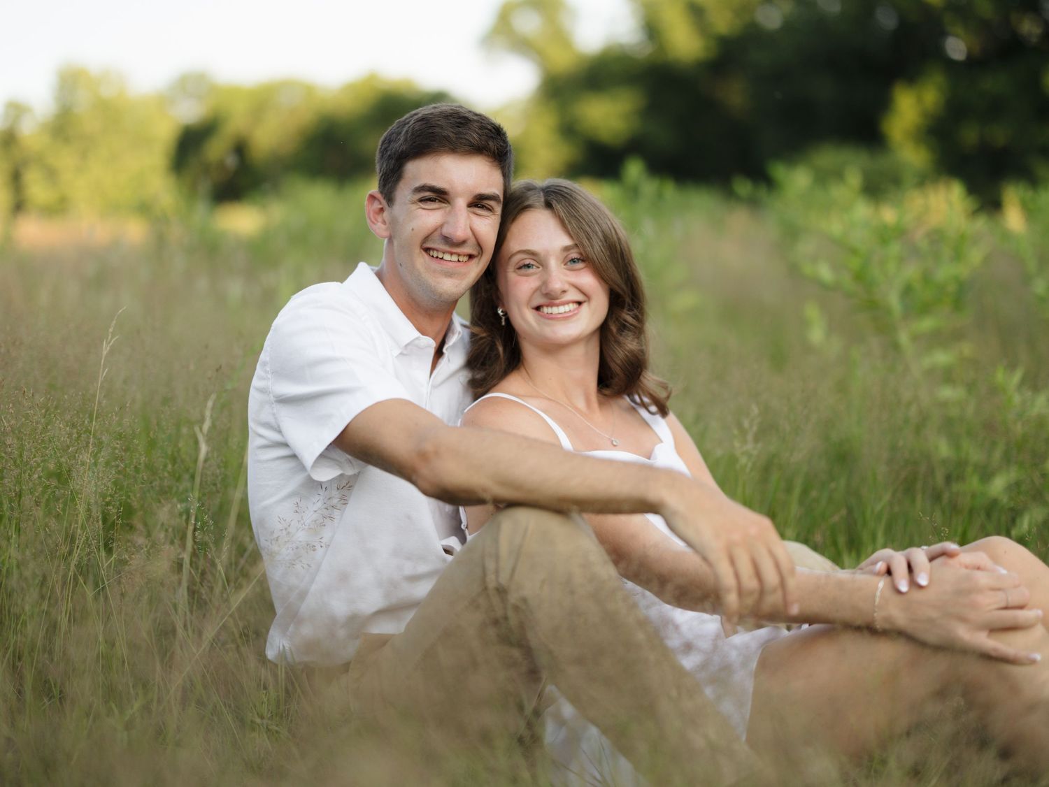Smiling young couple sitting close together in a tall grassy field, wearing casual summer clothes on a warm sunny day.