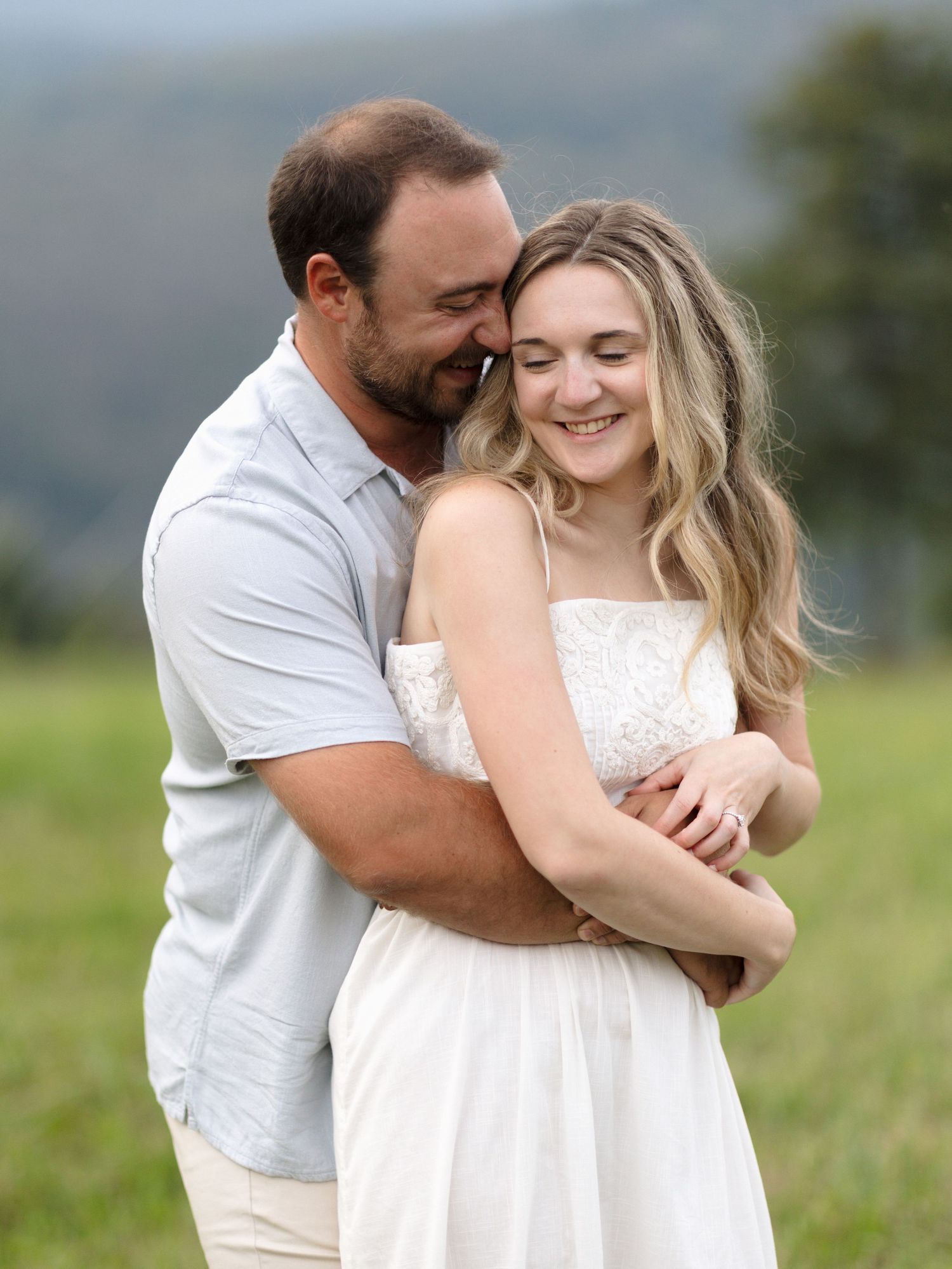 A smiling woman in a white lace dress is hugged from behind by a bearded man in a grassy field with hills behind.