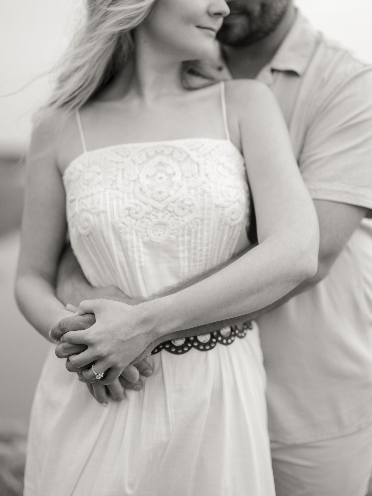 Black and white photo of a man embracing a woman in a white lace sundress from behind, their hands clasped at her waist.