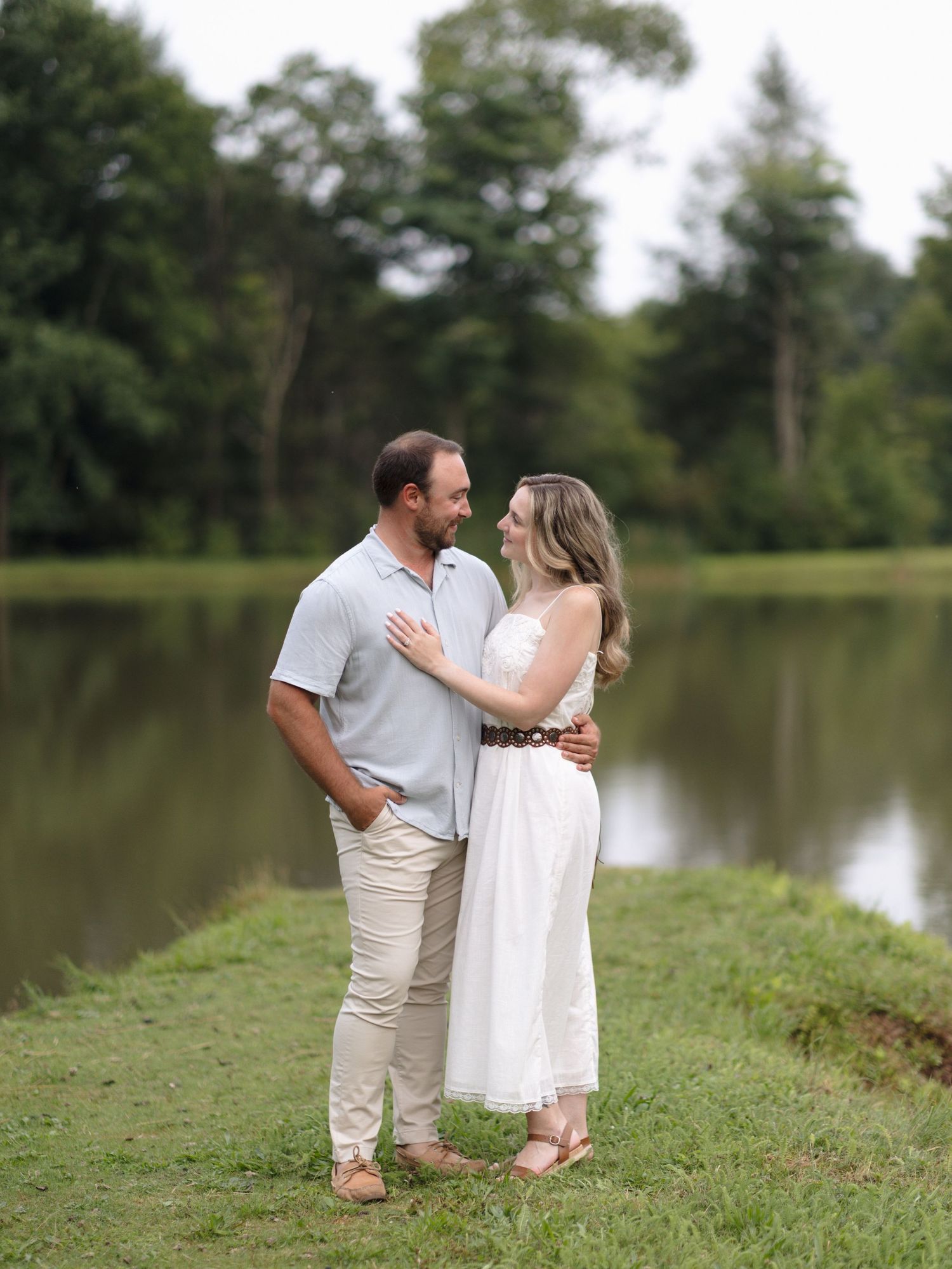 Smiling couple embracing on a grassy bank beside a calm pond, with lush green trees behind, in a romantic outdoor engagement-style portrait.