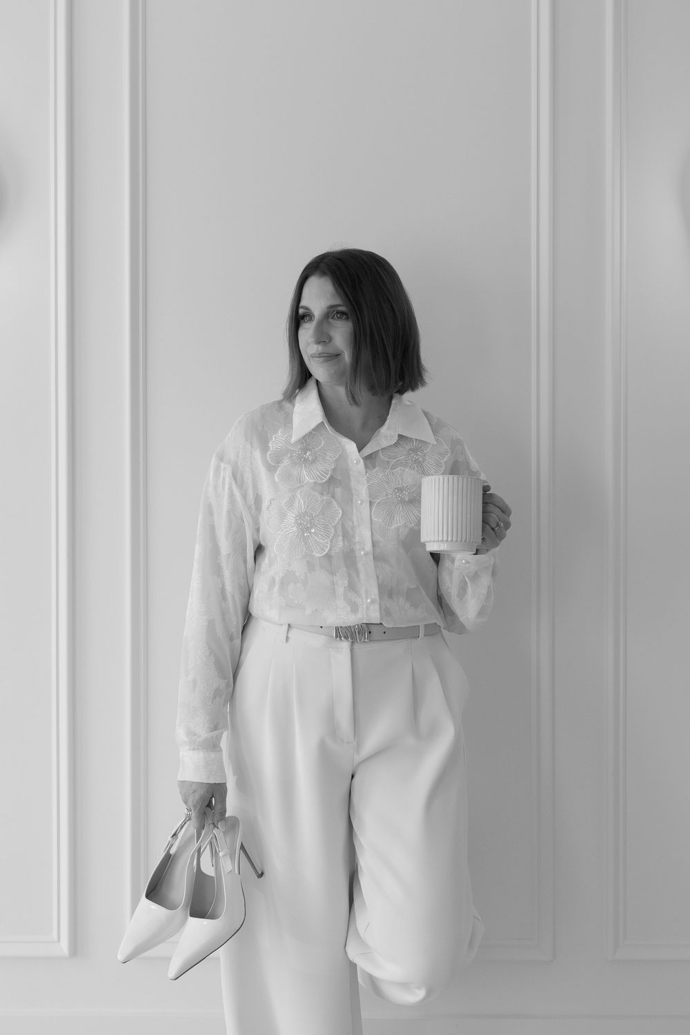 Black and white portrait of woman in white blouse and trousers holding a mug and heels against wall.