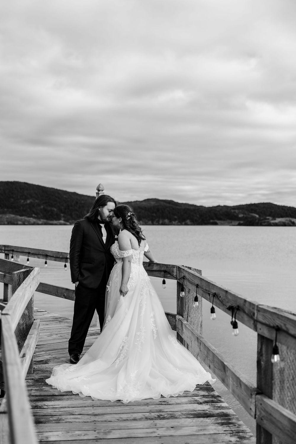Black and white photo of bride and groom sharing a romantic moment on a wooden pier overlooking a scenic lake.