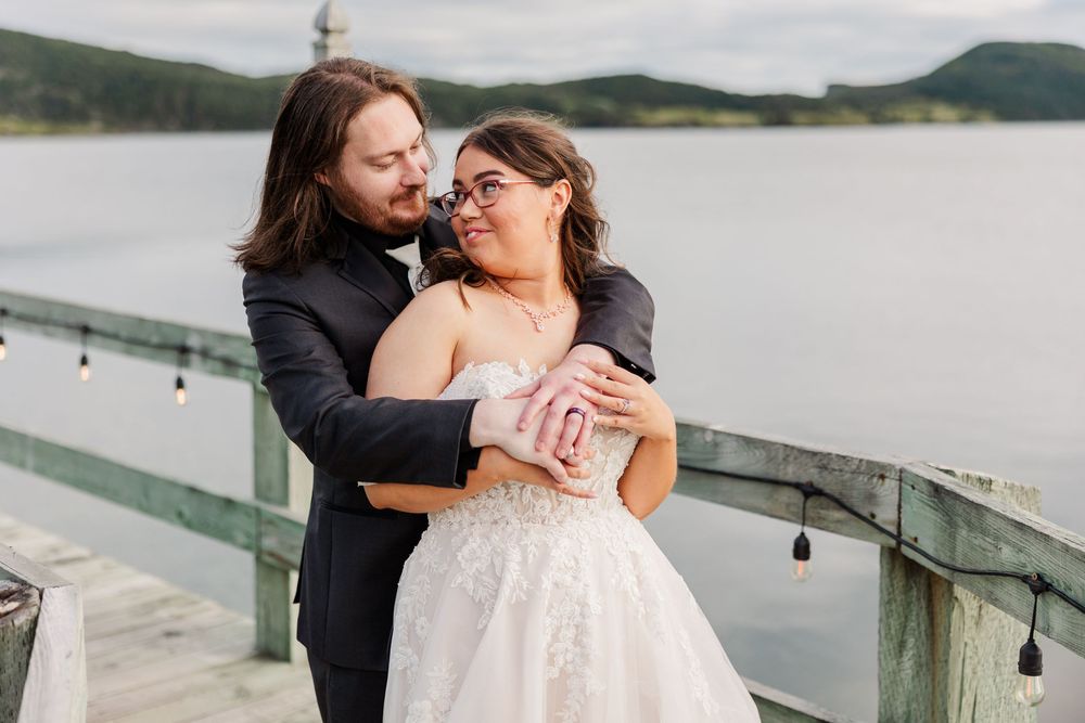 Bride and groom embrace on a waterfront dock, she in a white lace gown, he in a dark suit, smiling warmly.