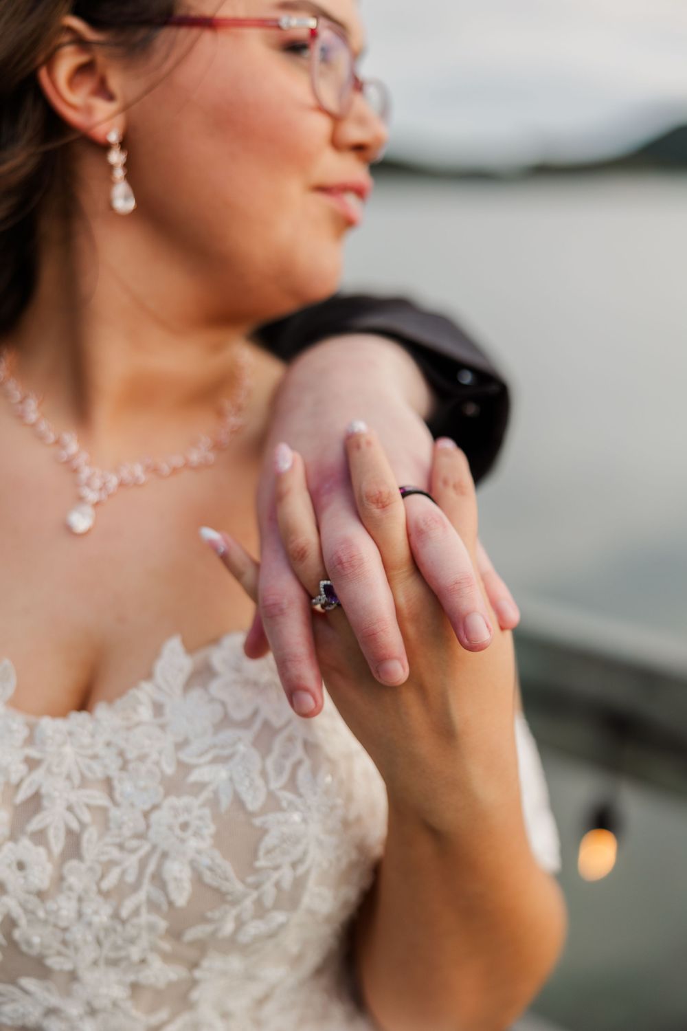 Bride in lace wedding dress showing off engagement ring and wedding band, wearing pearl jewelry and glasses.
