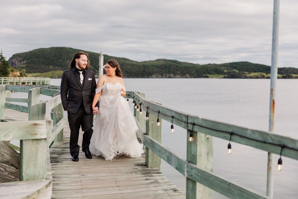 Bride and groom walking hand in hand on a wooden pier overlooking a scenic lake and green hills.