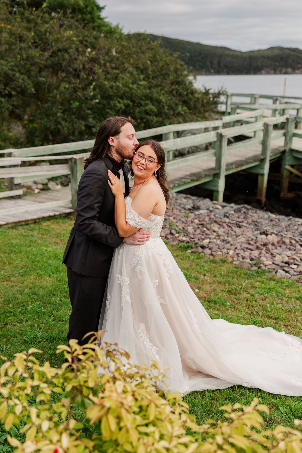Groom in black suit kisses bride wearing white lace ball gown near wooden dock with water and green trees.