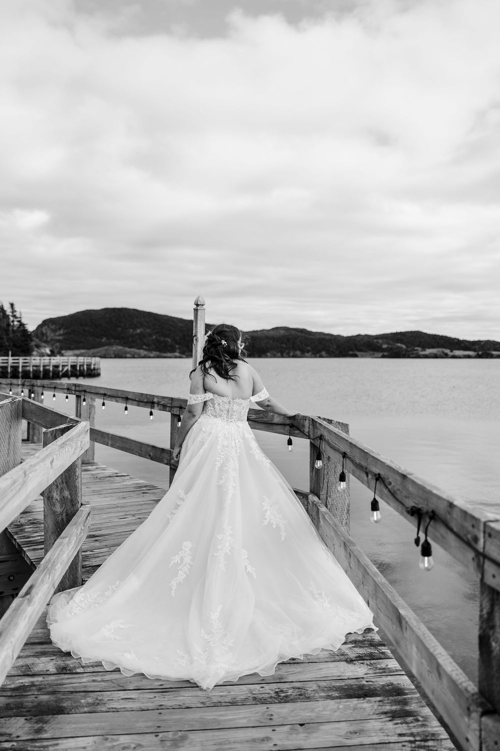 Black and white photo of a bride in a flowing lace gown standing on a wooden pier overlooking a serene lake.