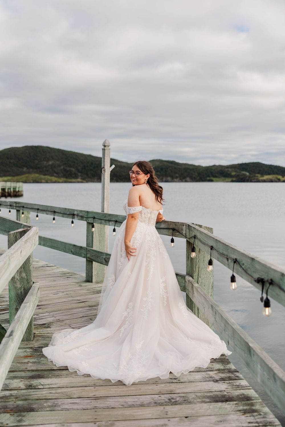 Bride in off-shoulder lace gown stands on wooden pier overlooking calm water with hills in background.
