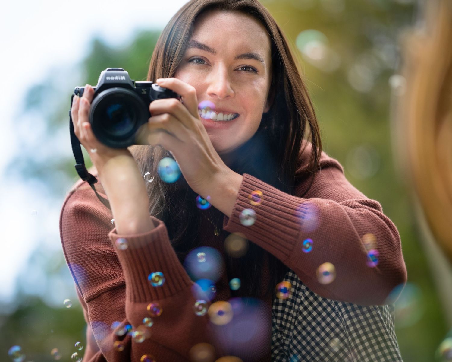 Smiling female photographer holding DSLR camera with colorful soap bubbles floating around her.