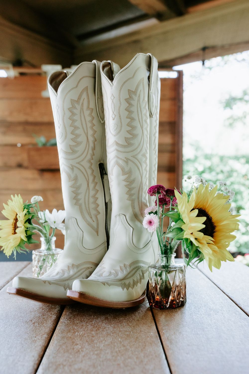 White embroidered cowboy boots on a wooden table beside sunflower and pink wildflower bouquets in glass vases.
