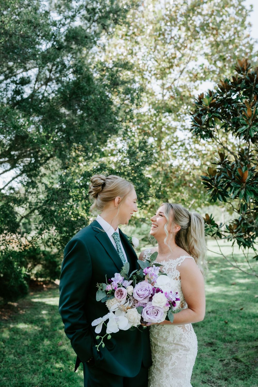 Two brides smile lovingly at each other outdoors, one in a dark green suit and one in a lace gown, holding a purple floral bouquet.