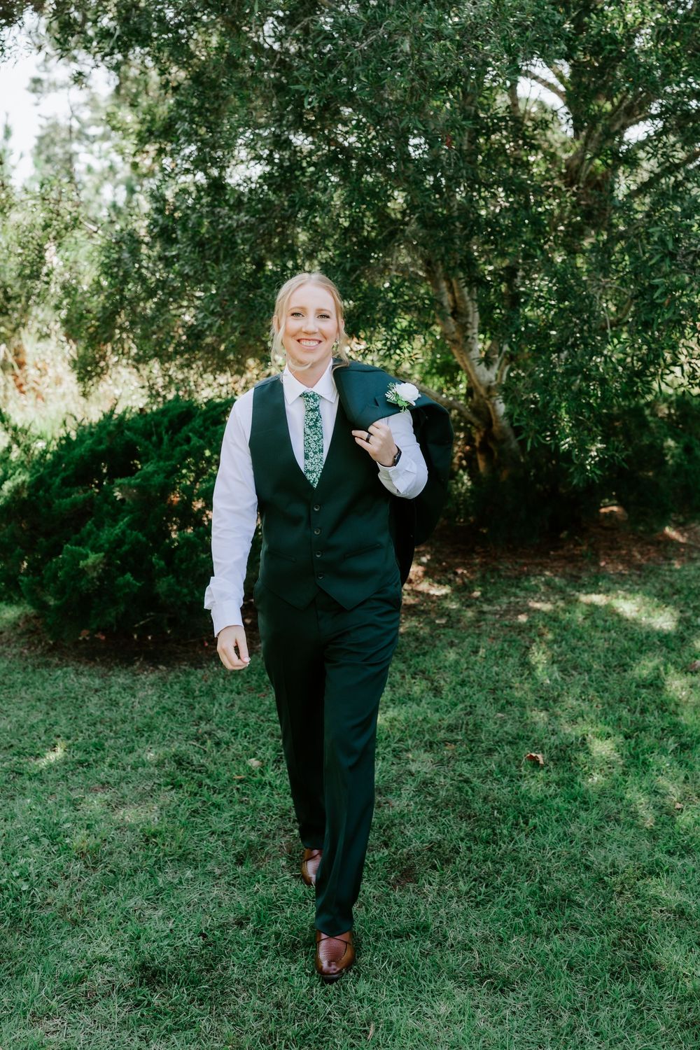 Smiling person in a dark green vest, white shirt and floral tie walks across a grassy lawn with a jacket slung over one shoulder.