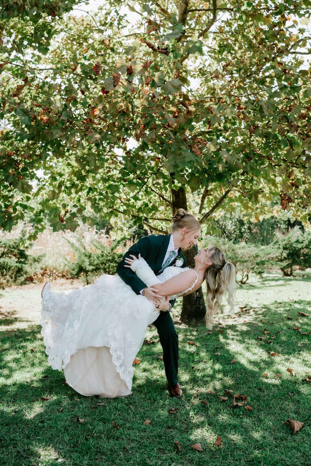 Groom in a dark suit dips his bride in a white lace wedding gown beneath a leafy autumn tree on a sunlit lawn.