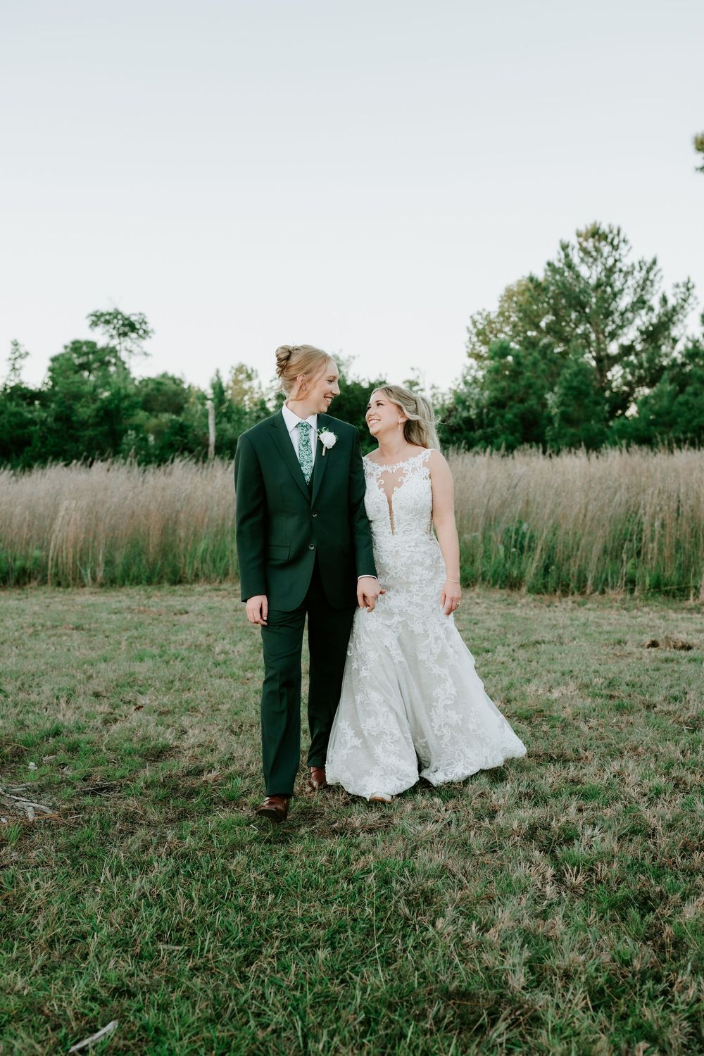 Bride in a lace wedding gown and groom in a green suit hold hands and smile at each other in a grassy field at sunset.