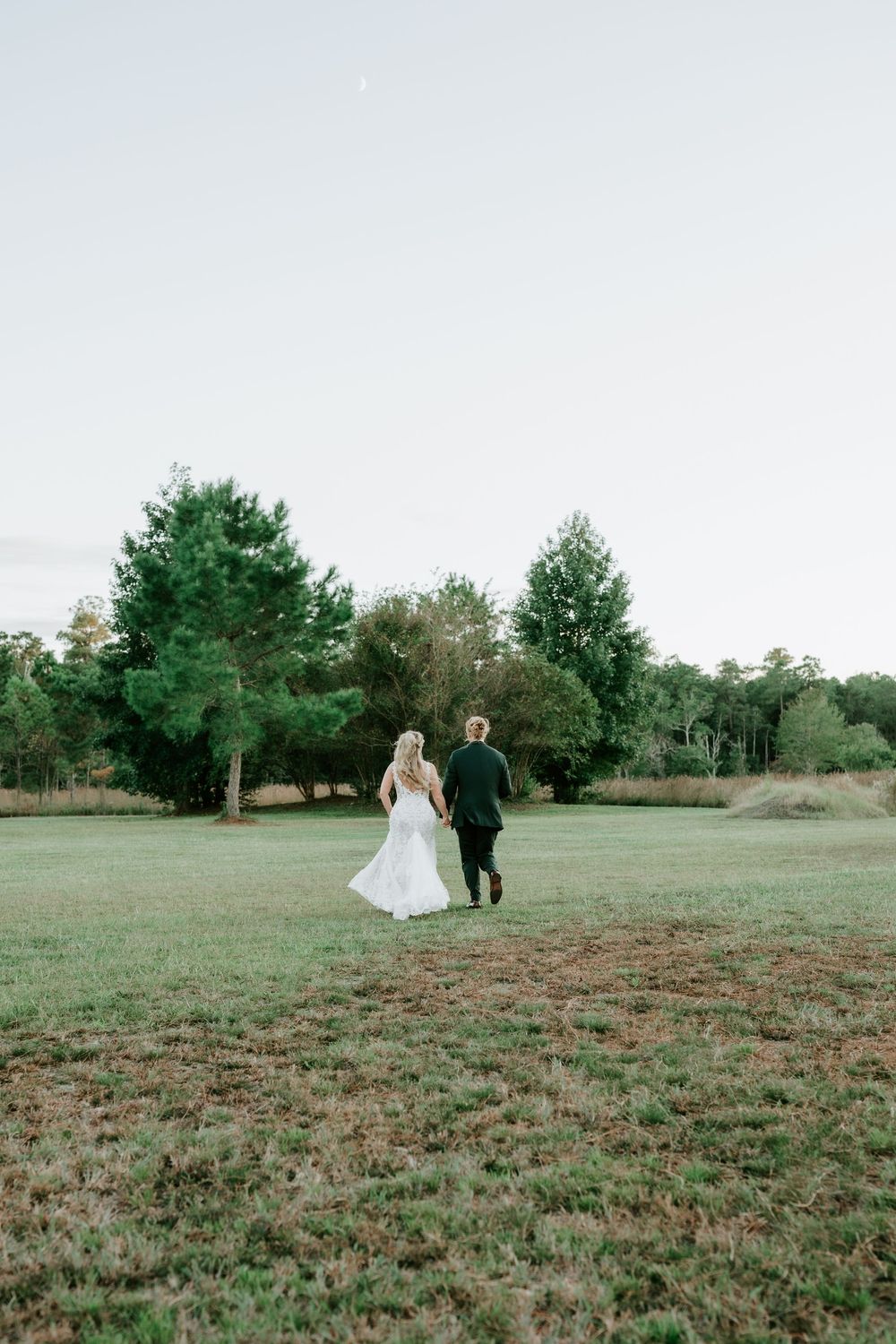 Bride in a white gown and groom in a dark suit hold hands and walk away across a grassy field toward trees at dusk.