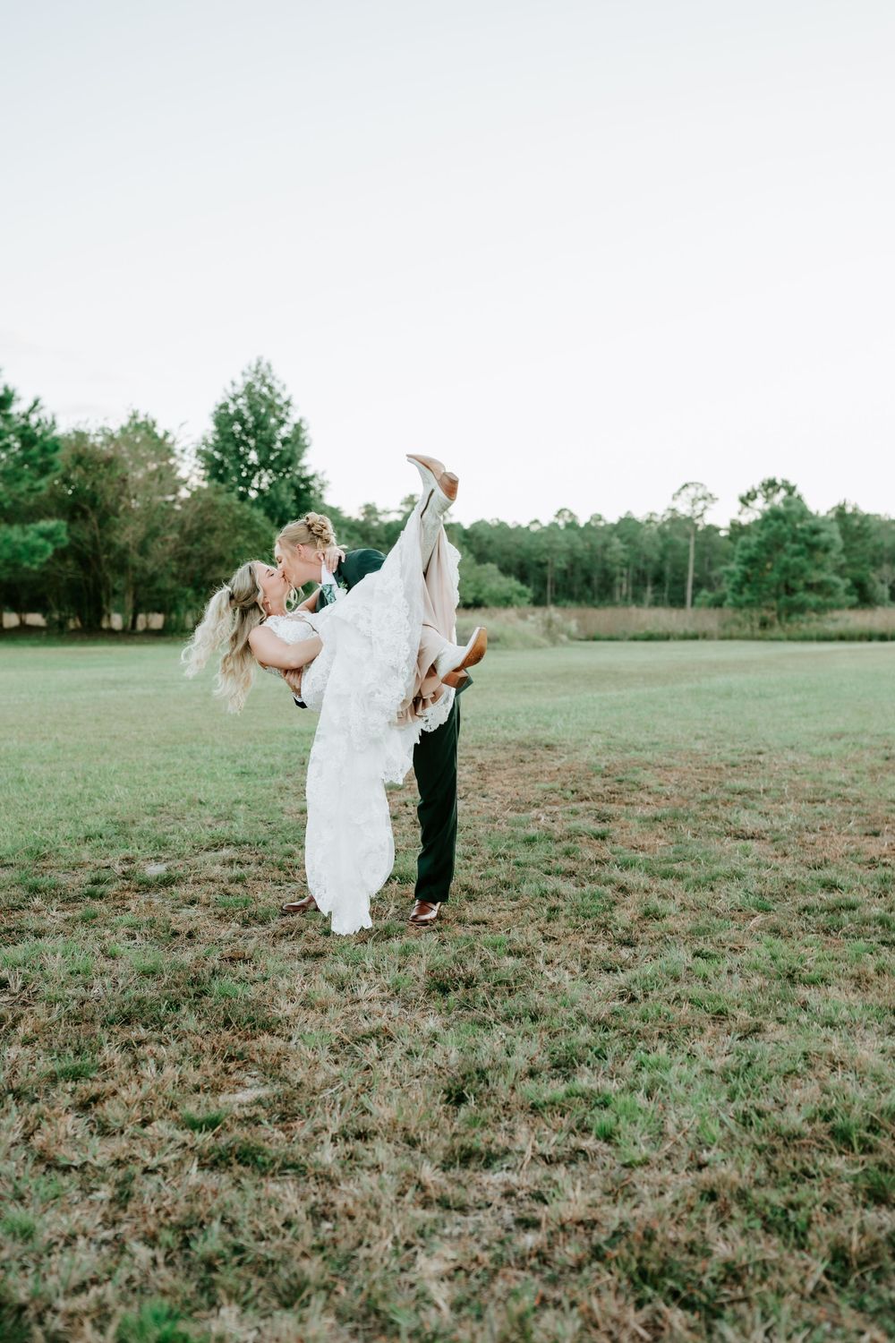 A groom dips a bride in a flowing lace wedding gown for a kiss in a grassy field with tall trees behind them.