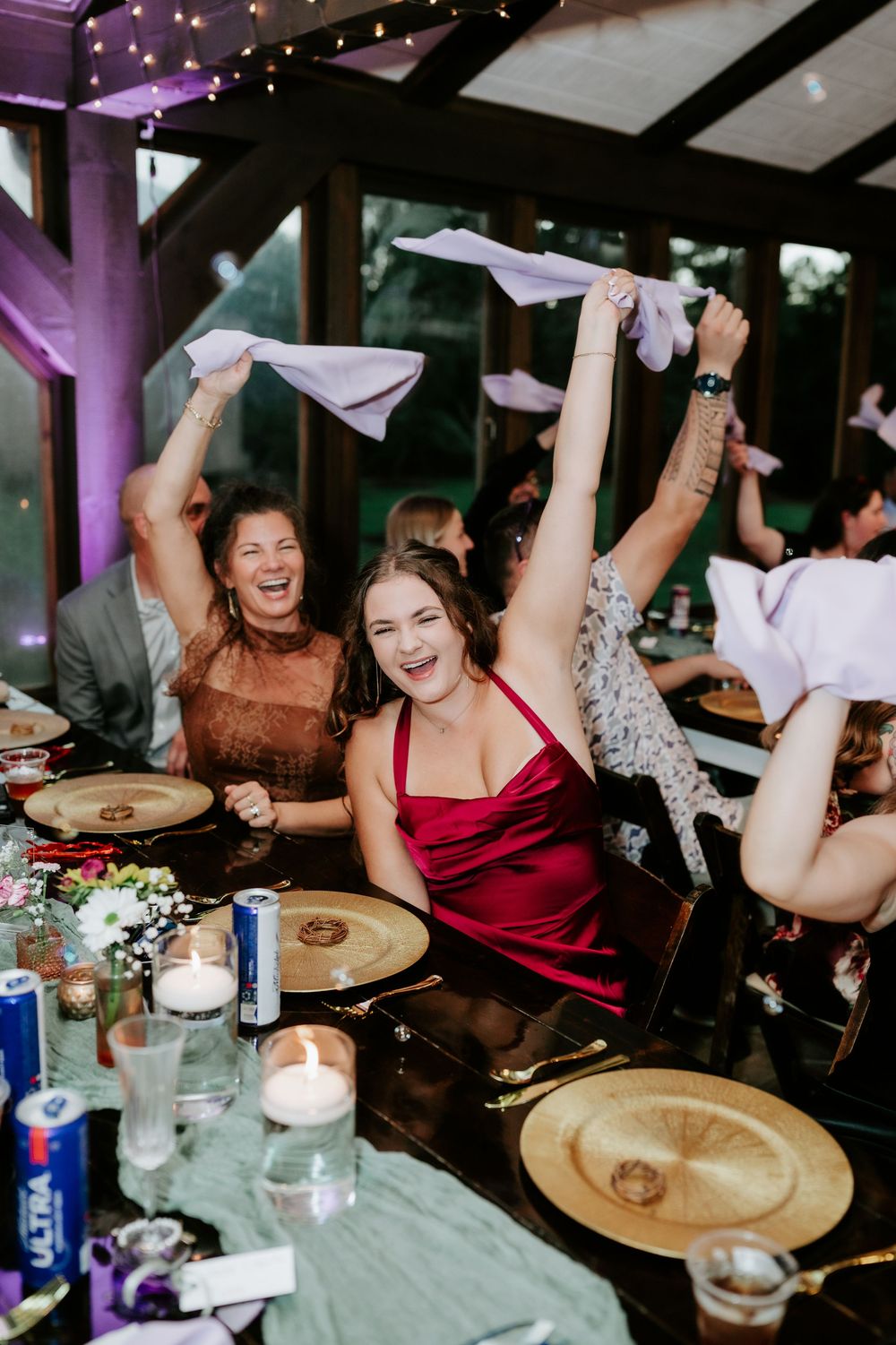 Smiling wedding guests in a rustic venue wave white napkins in the air while seated at a candlelit table with gold charger plates.