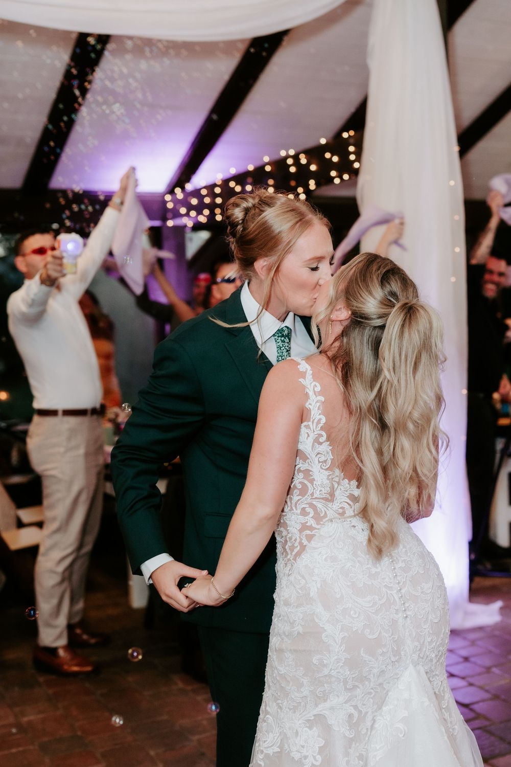 Bride in a lace gown and groom in a green suit share a kiss on the dance floor as wedding guests celebrate under twinkling string lights.