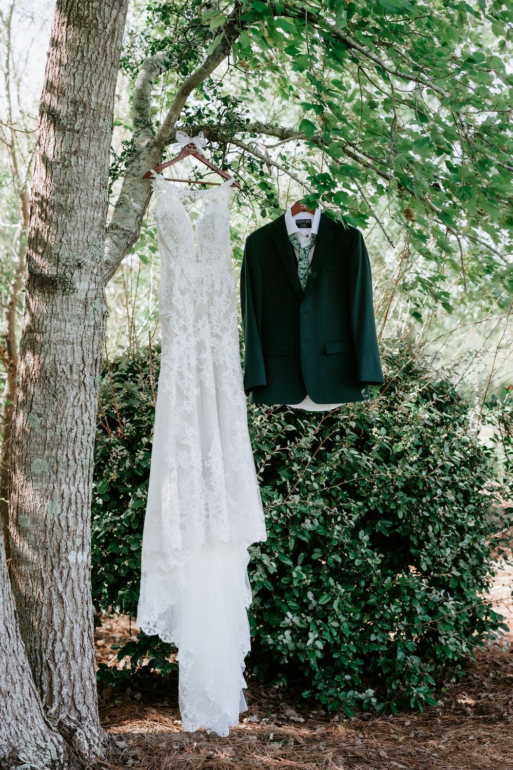 Lace wedding gown and dark green groom's suit jacket hanging from tree branches outdoors, framed by lush green foliage.