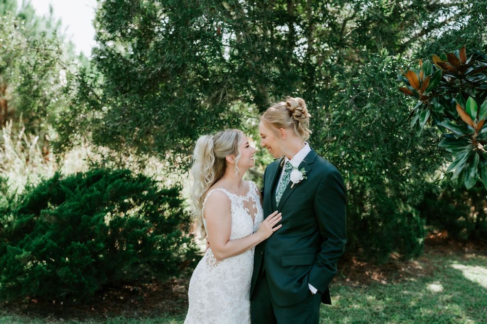 Bride in a white lace wedding dress and groom in a dark suit with a boutonniere share a close moment outdoors surrounded by green trees.
