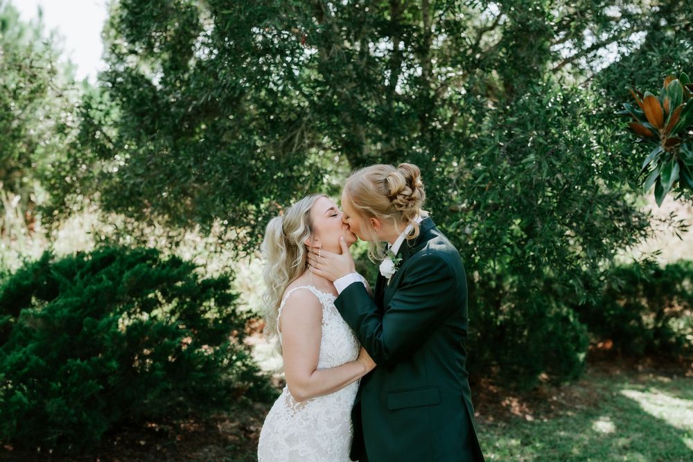Bride in a lace wedding dress and groom in a dark suit share a kiss outdoors in a sunlit garden surrounded by lush green trees.