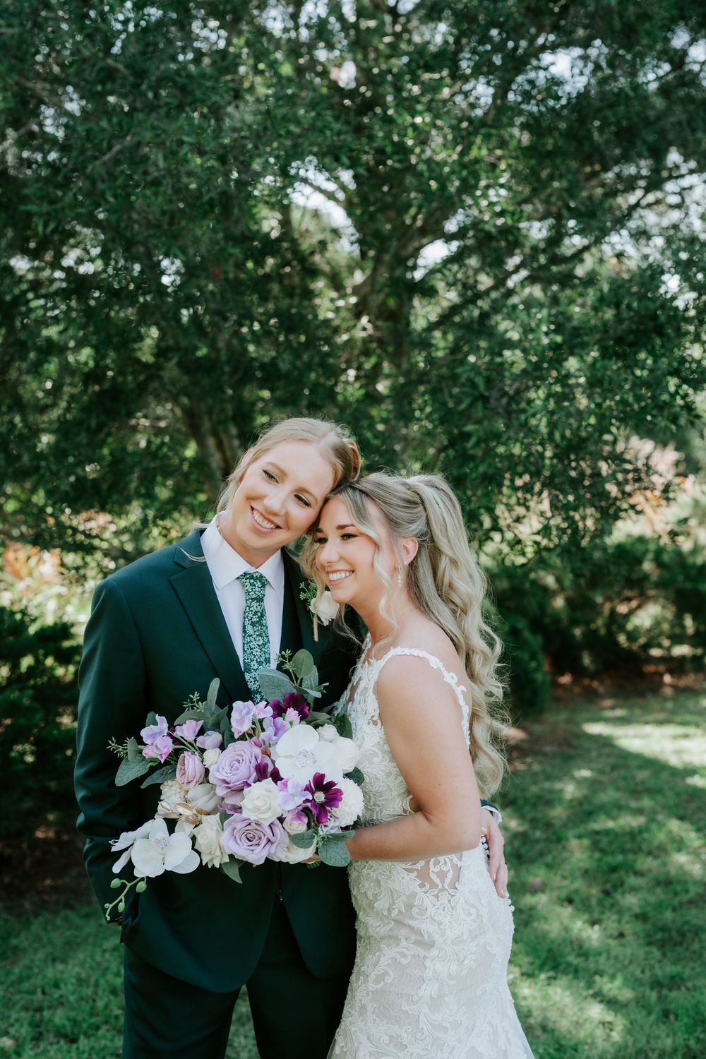 Smiling bride in a lace wedding gown and groom in a dark green suit embrace outdoors holding a purple and white bouquet under leafy trees.
