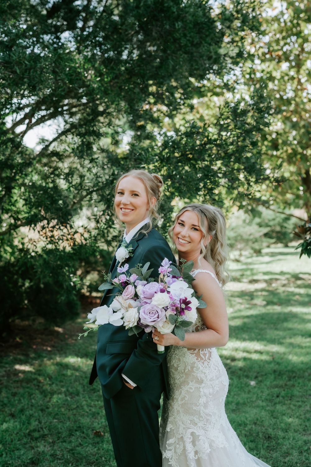 Two smiling brides embrace outdoors under green trees, one in a dark green suit and one in a lace gown holding a purple and white bouquet.