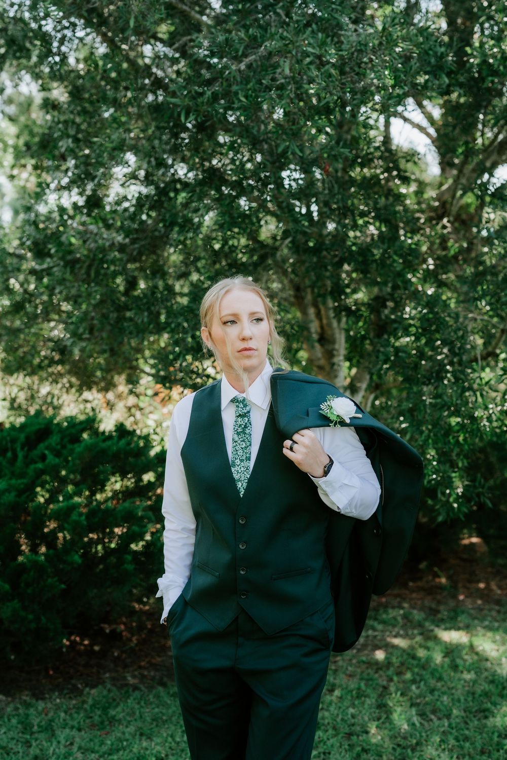 A person in a dark vest, white shirt and green patterned tie holds a jacket over one shoulder beside leafy green trees.