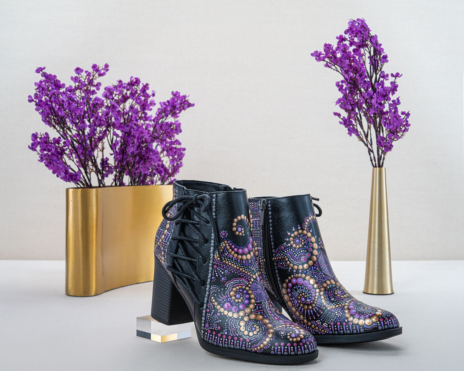 Black lace-up ankle boots with hand-painted purple and gold dot mandala designs beside gold vases holding purple flowers.