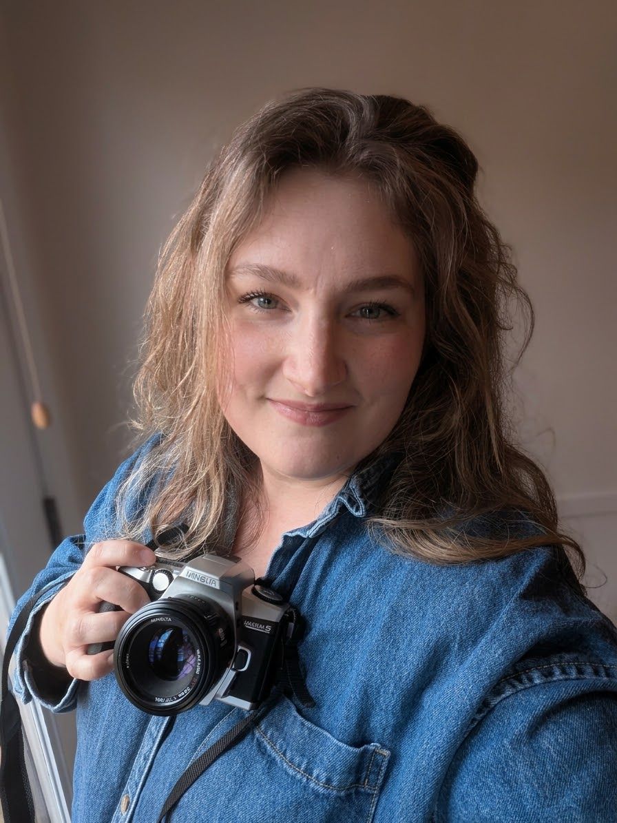 Black and white portrait of a smiling woman holding a vintage film camera, with curly hair and a denim jacket.
