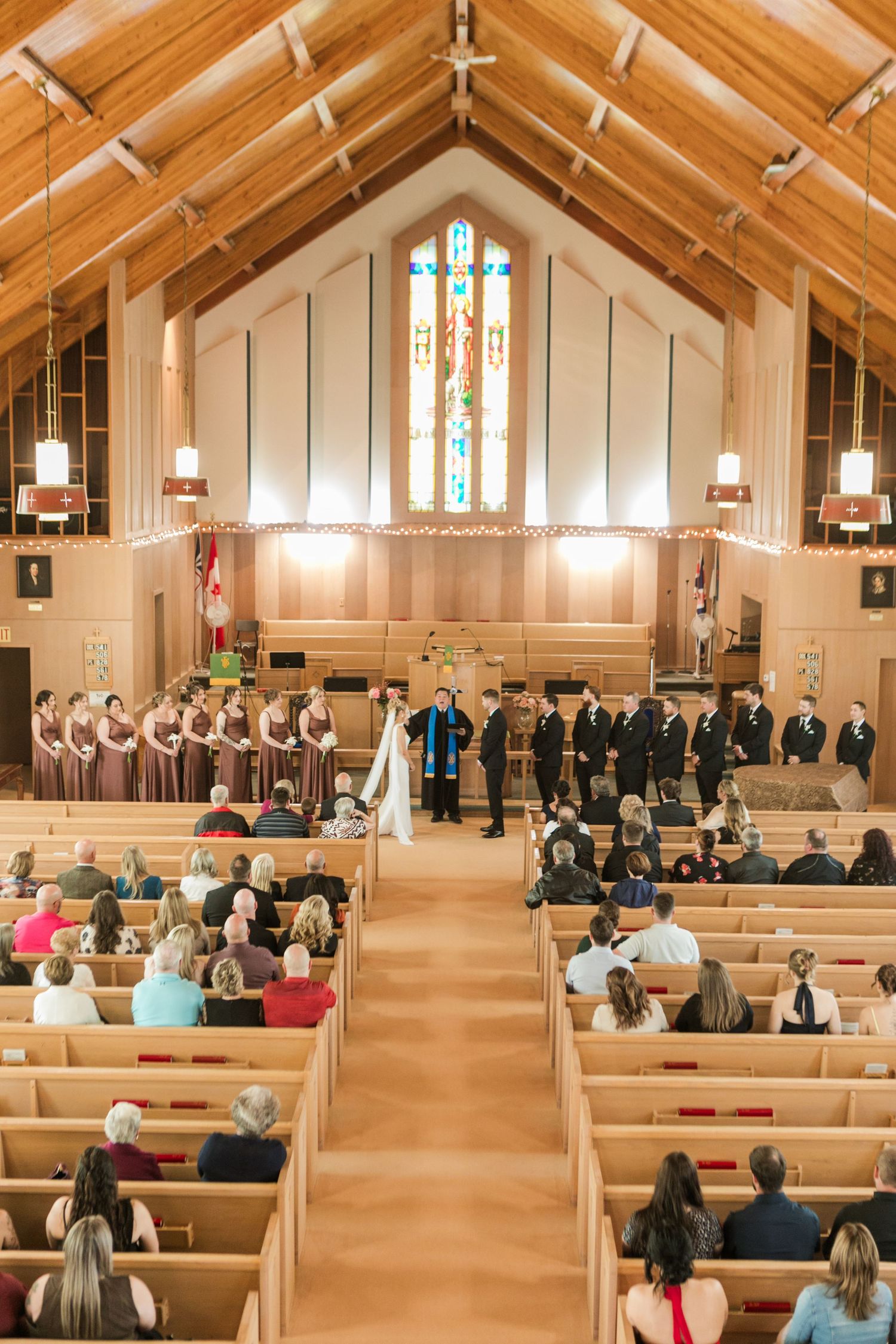 Wedding ceremony in a wooden church with stained glass, bride and groom at altar, bridesmaids and groomsmen, guests in pews.