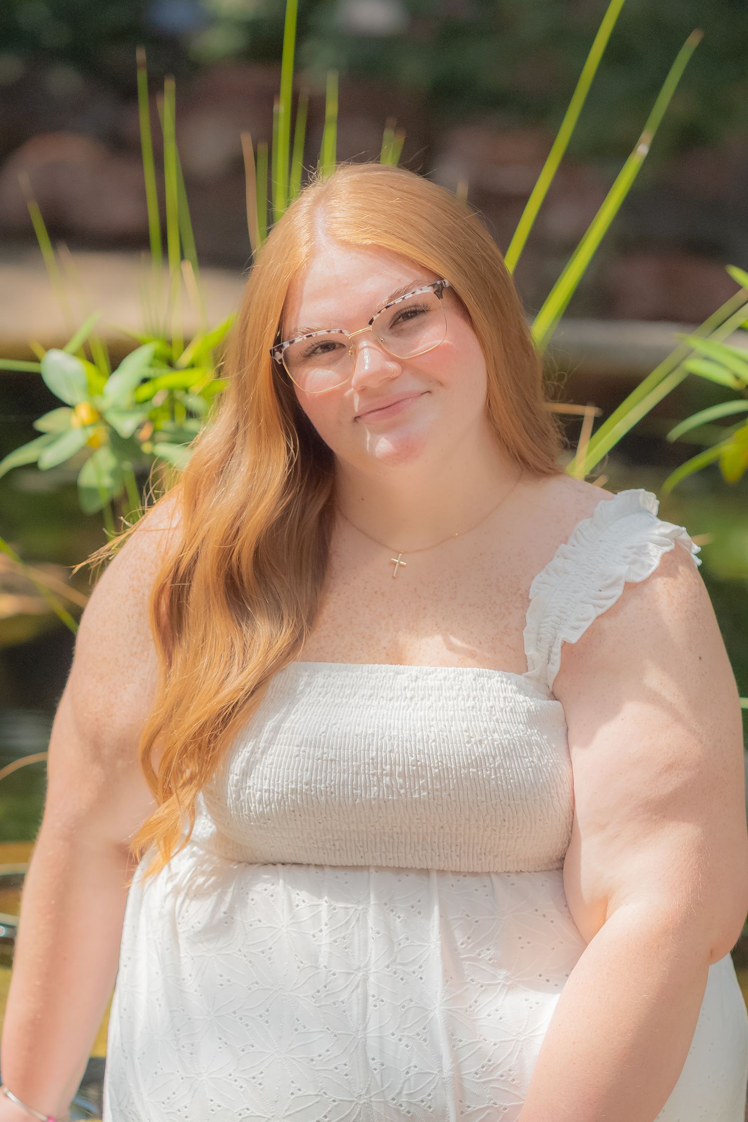 Young woman with long red hair and glasses in white dress smiling outdoors near green plants.