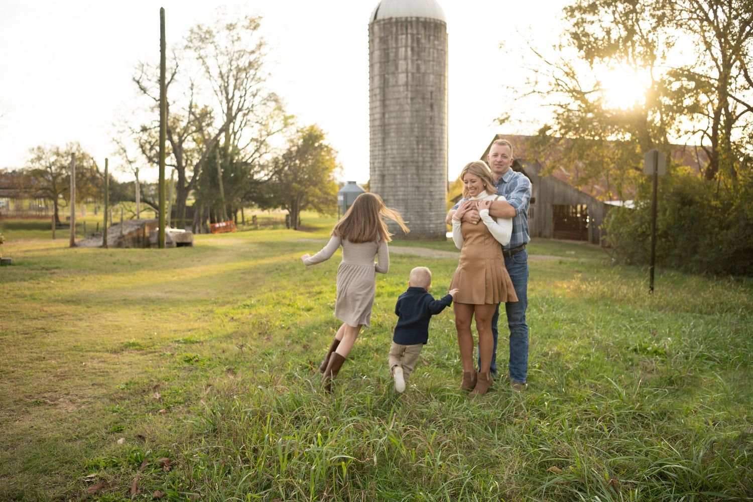 Family playing outdoors near rustic barn, parents holding white fabric while toddler runs through, fall family photo session.