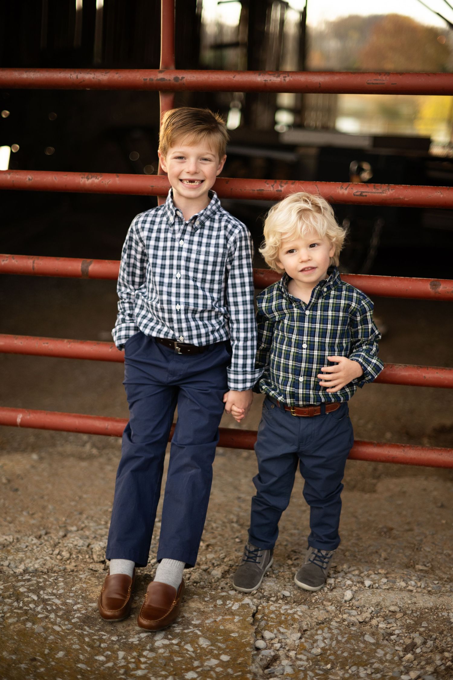 Two young boys in matching plaid shirts and navy pants pose together by a red metal fence outdoors.