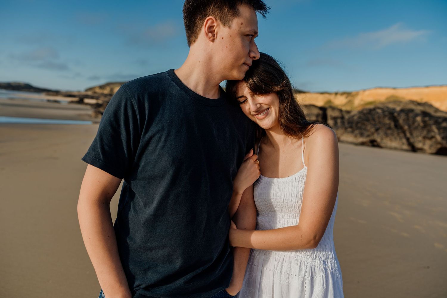 Couple holding hands barefoot on wet sandy beach, woman in white dress and man in jeans at golden hour.