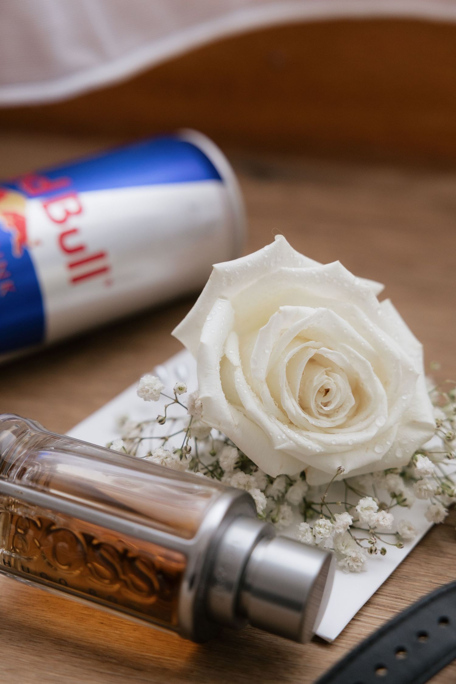 White rose boutonniere with baby's breath beside Hugo Boss cologne and Red Bull can on wooden surface.