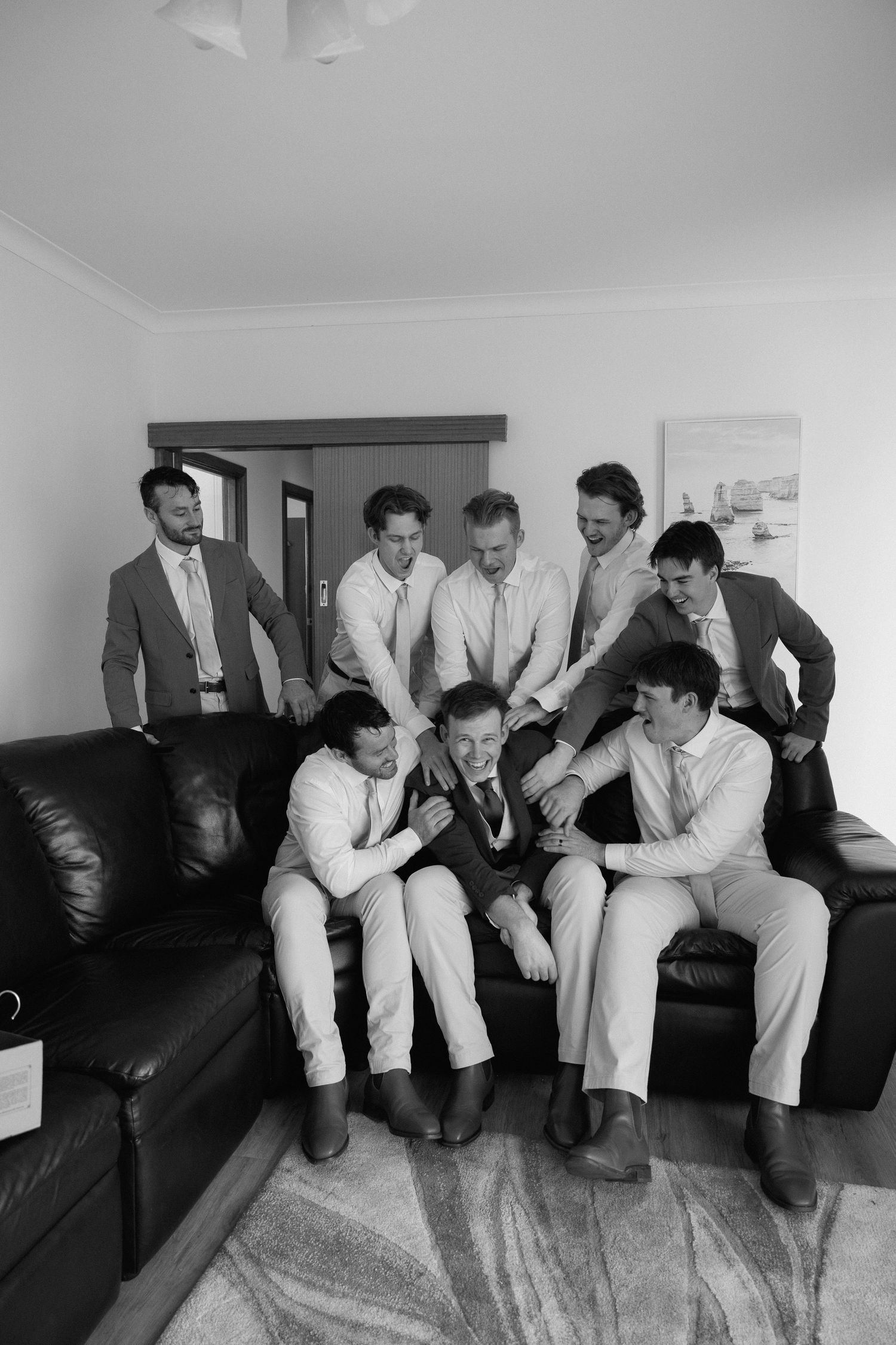 Groomsmen laughing and gathered around groom on leather sofa during wedding preparations in black and white.