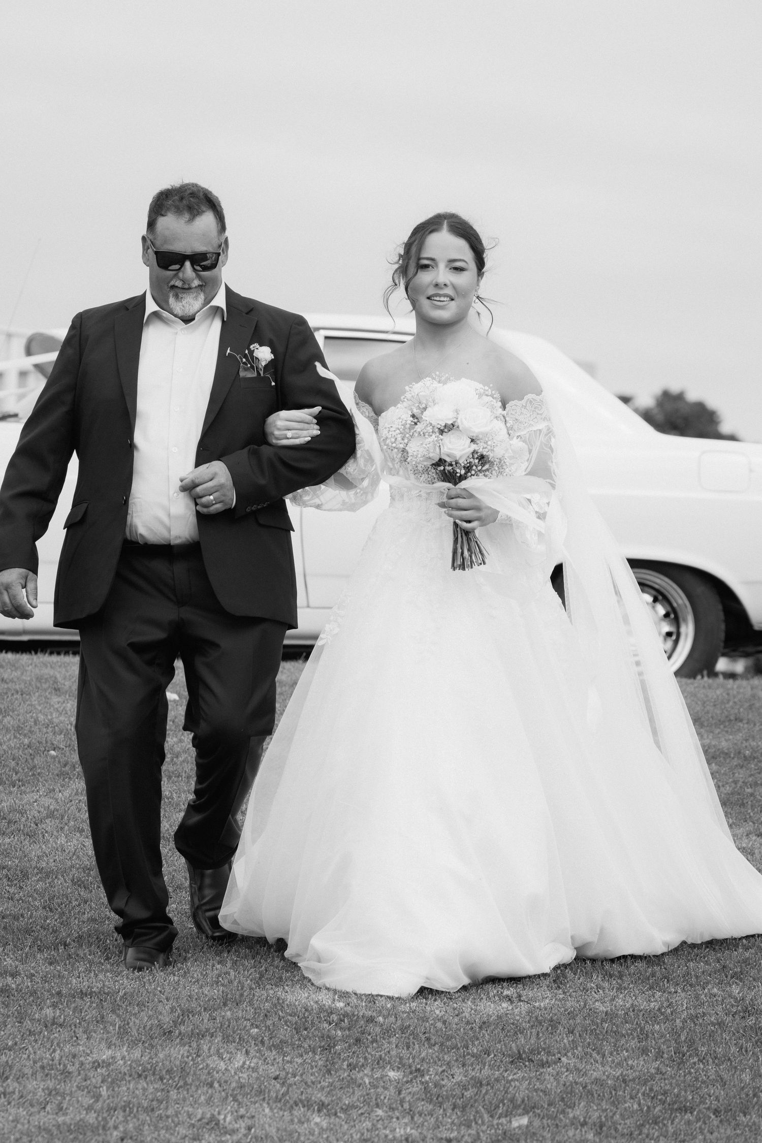 Black and white photo of a smiling bride in a white gown holding a bouquet, walking arm in arm with a suited man.