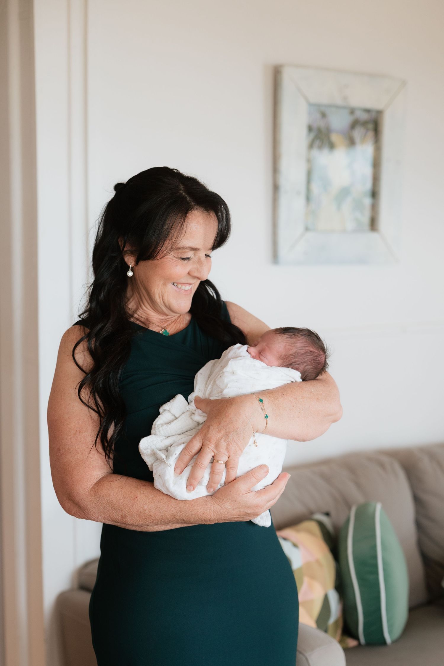A smiling woman with dark hair in a green dress tenderly holds a swaddled newborn baby indoors.