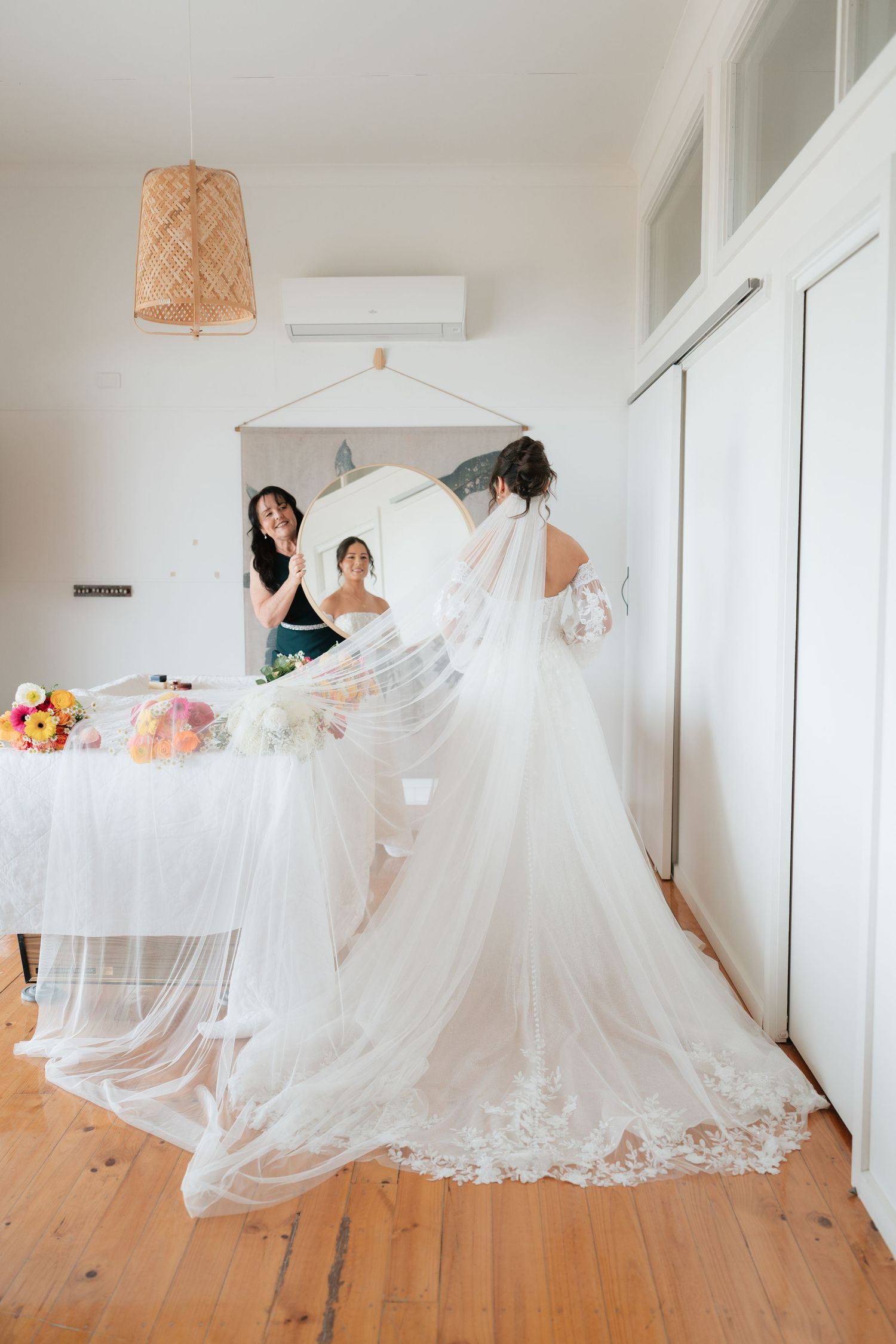 Bride in a flowing lace wedding gown with veil admires her reflection in a mirror while a bridesmaid assists.